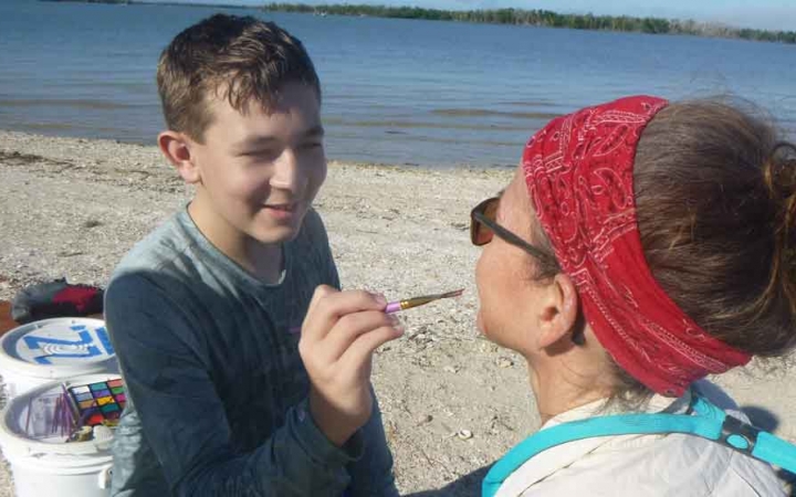a young person paints on their parent's face on an outward bound expedition for families. They appear to be on a beach.